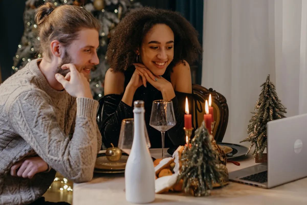 A couple at a candlelit Christmas table smiling at an open laptop during a video call