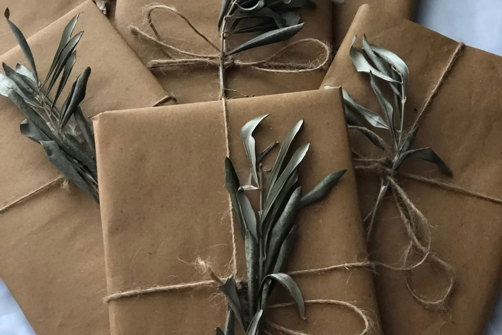 Flat parcels wrapped in brown paper and tied with twine, each with a sprig of olive leaves, on a white cloth