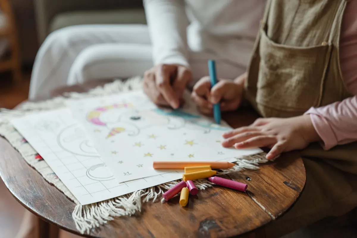 A child colours in a picture with a blue crayon at a small wooden table, an adult's hand beside theirs