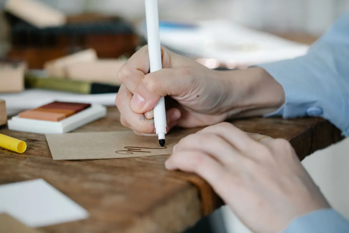 A hand writing on a small brown card with a white marker at a wooden table