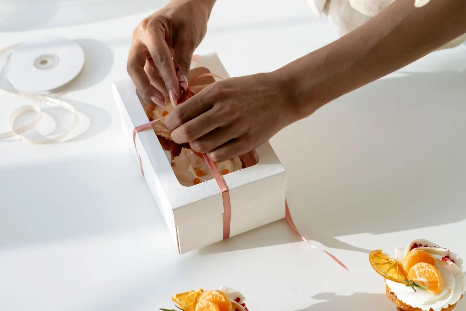 Hands tie a pink ribbon around a white bakery box of cakes on a sunlit white table