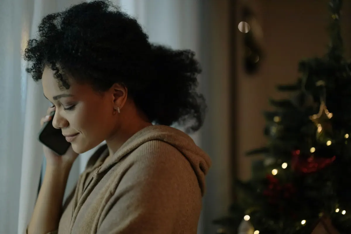 A woman in a warm hooded jumper smiles as she talks on a mobile phone, with a lit Christmas tree behind her