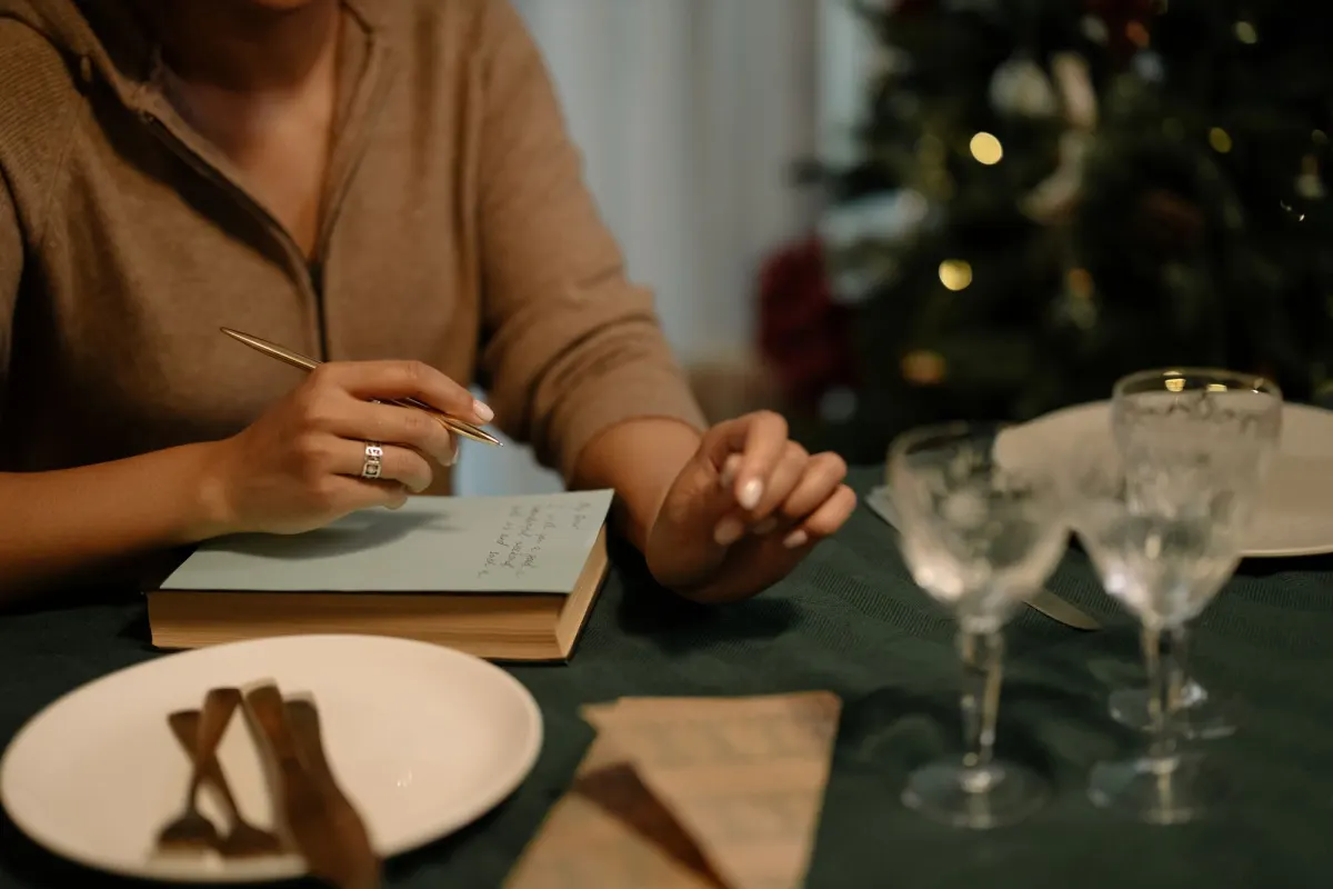 A woman writing a short note at a table set for Christmas dinner, with tree lights glowing behind her