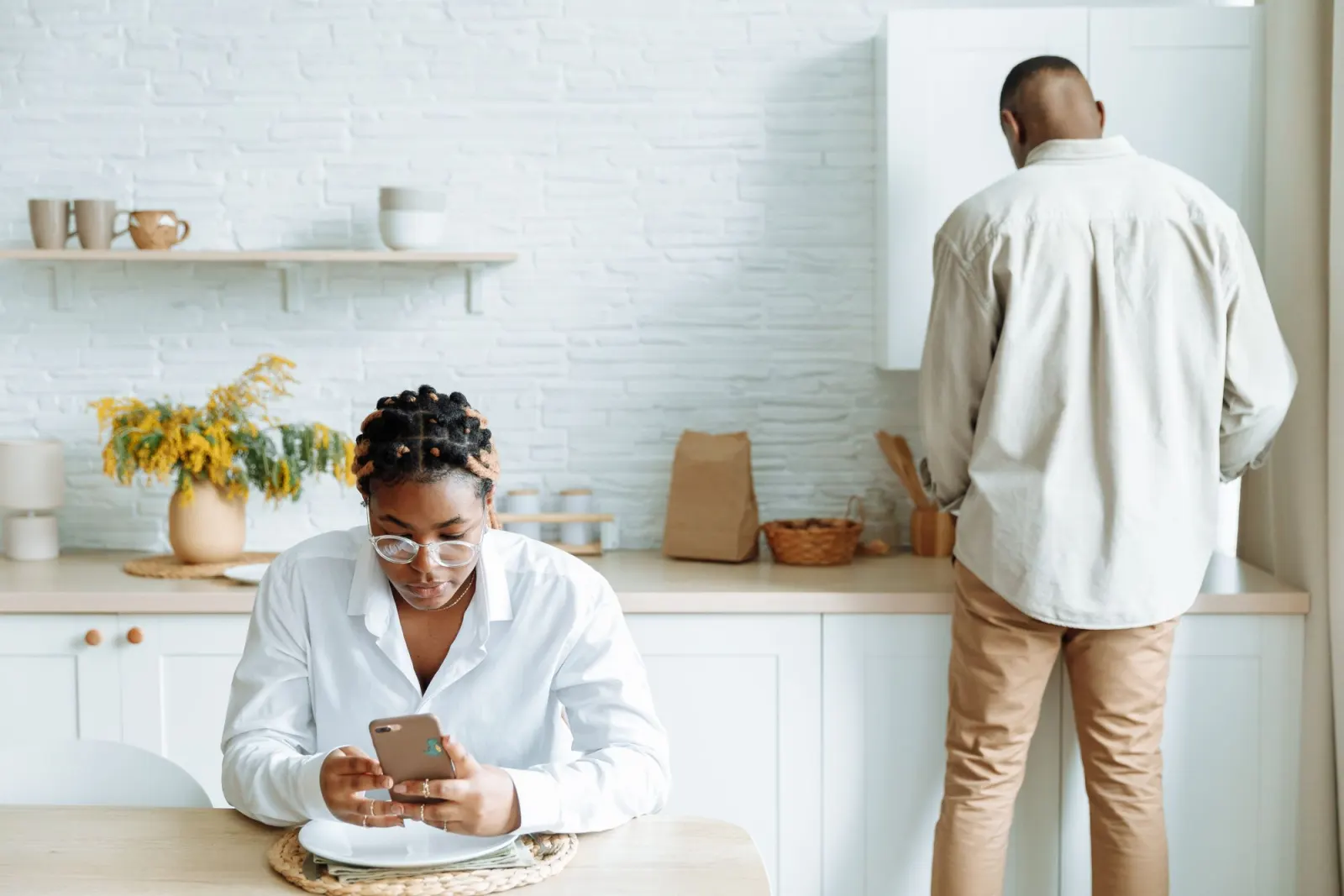 A woman at a kitchen table types on her phone while a man stands at the bench with his back to her