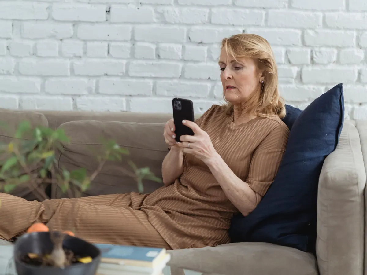 An older woman sits on a sofa with her feet up, looking closely at the phone in her hands
