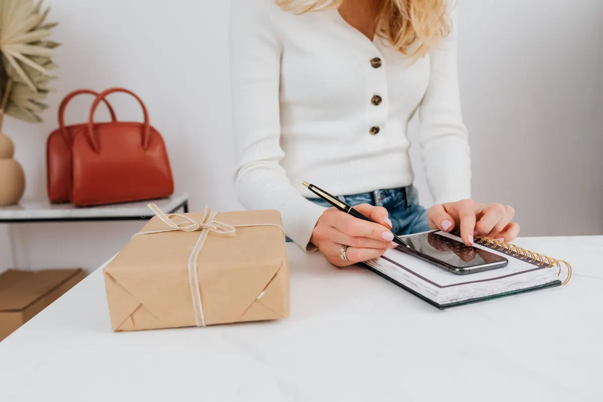 A woman's hands with a pen and a phone resting on a planner, next to a parcel wrapped in brown paper