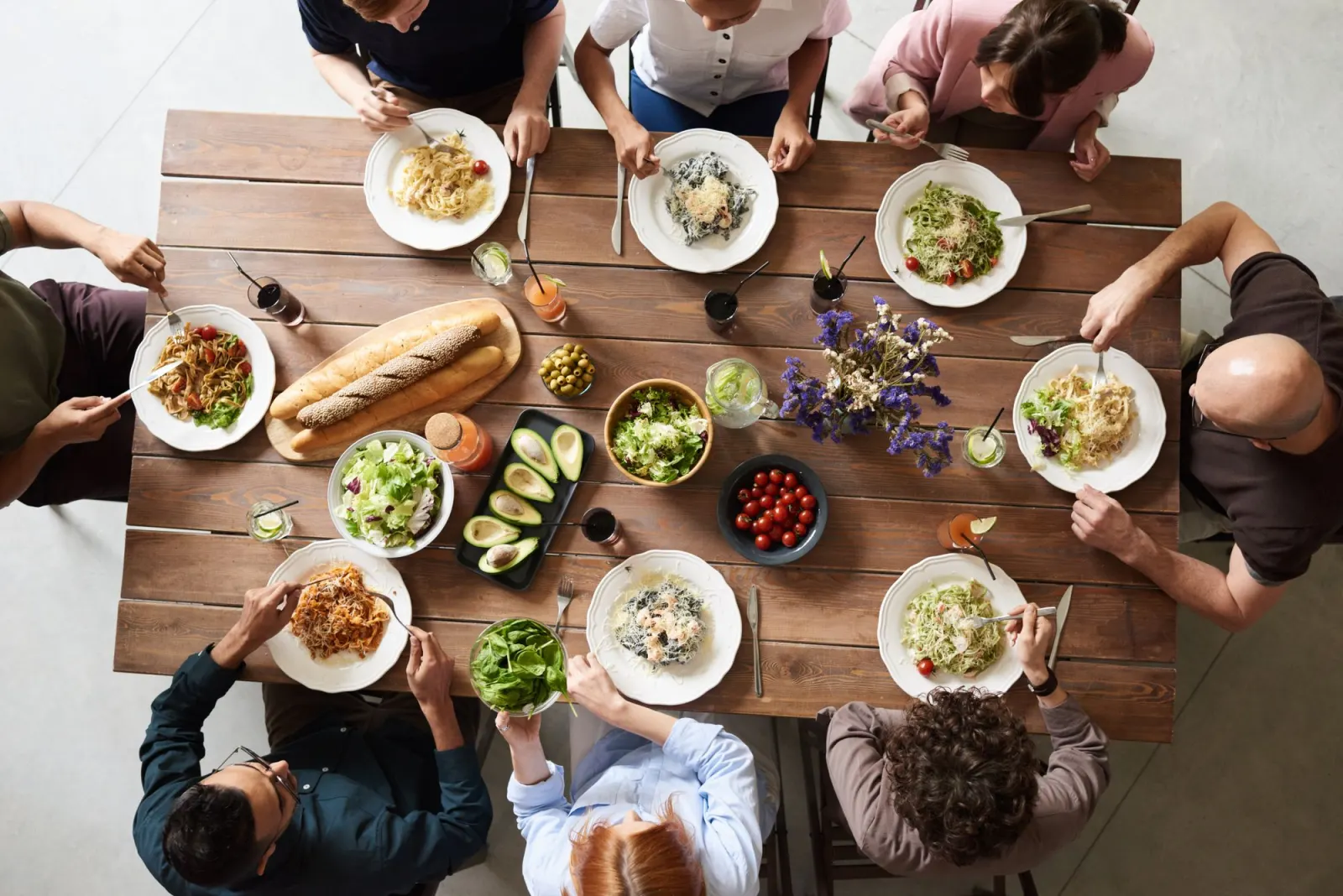 Overhead view of eight people sharing lunch at a long wooden table, with pasta, salads, bread and a vase of flowers