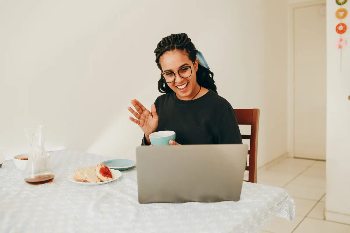 A woman holding a cup waves at her laptop during a video call at a kitchen table