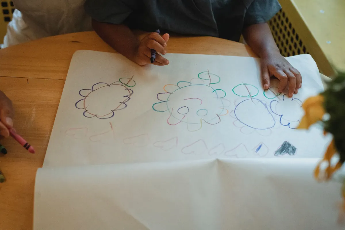 A child's hands drawing flowers and hearts with wax crayons on a large sheet of paper on a wooden table
