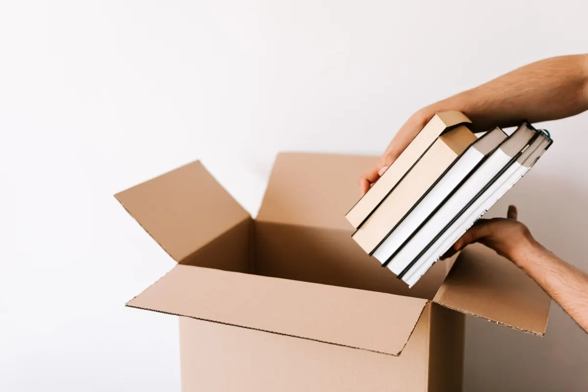Two hands lowering a stack of hardcover books into an open cardboard box against a white wall