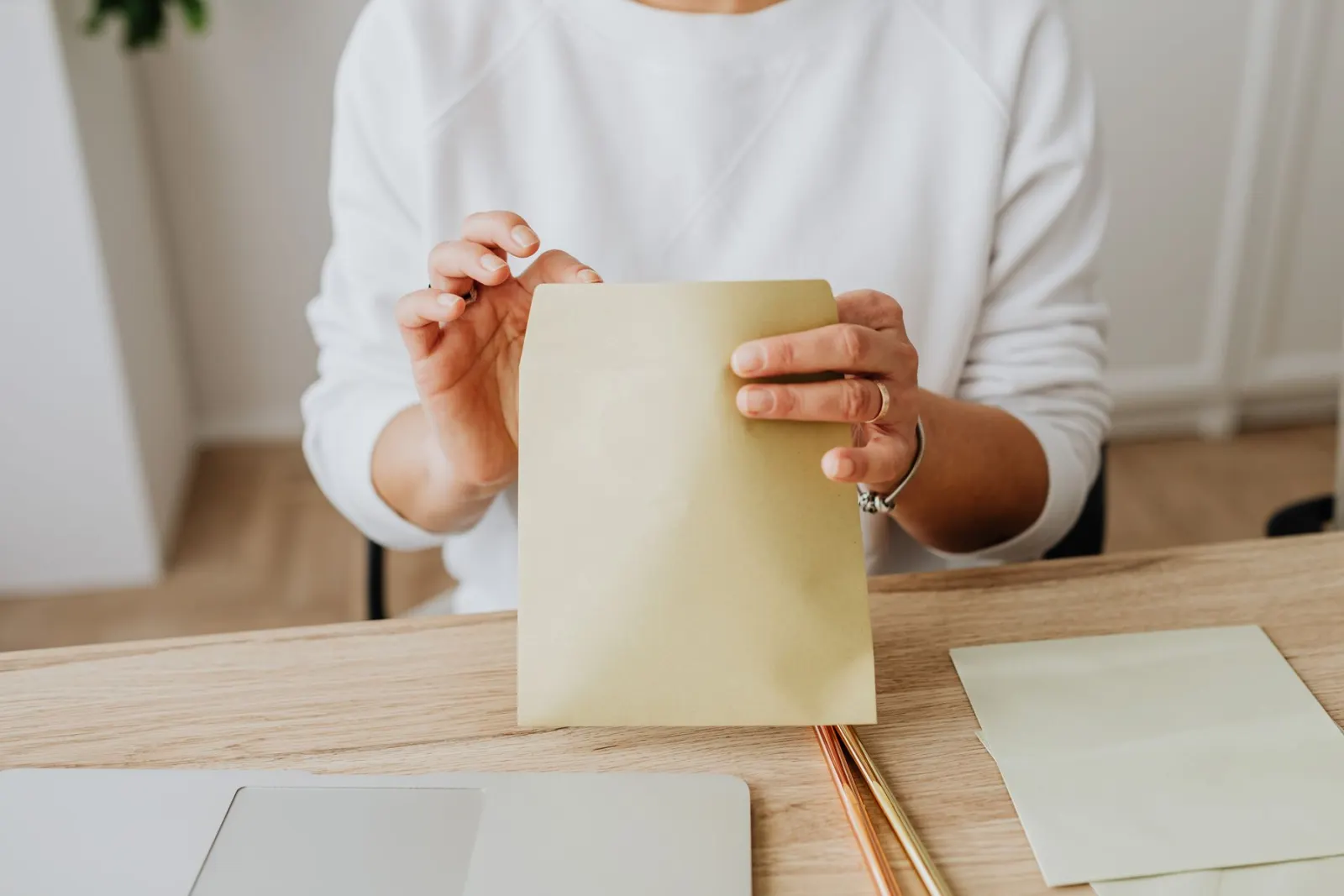 A woman at a desk beside a closed laptop holds a pale brown envelope, with blank cards and two pens in front of her