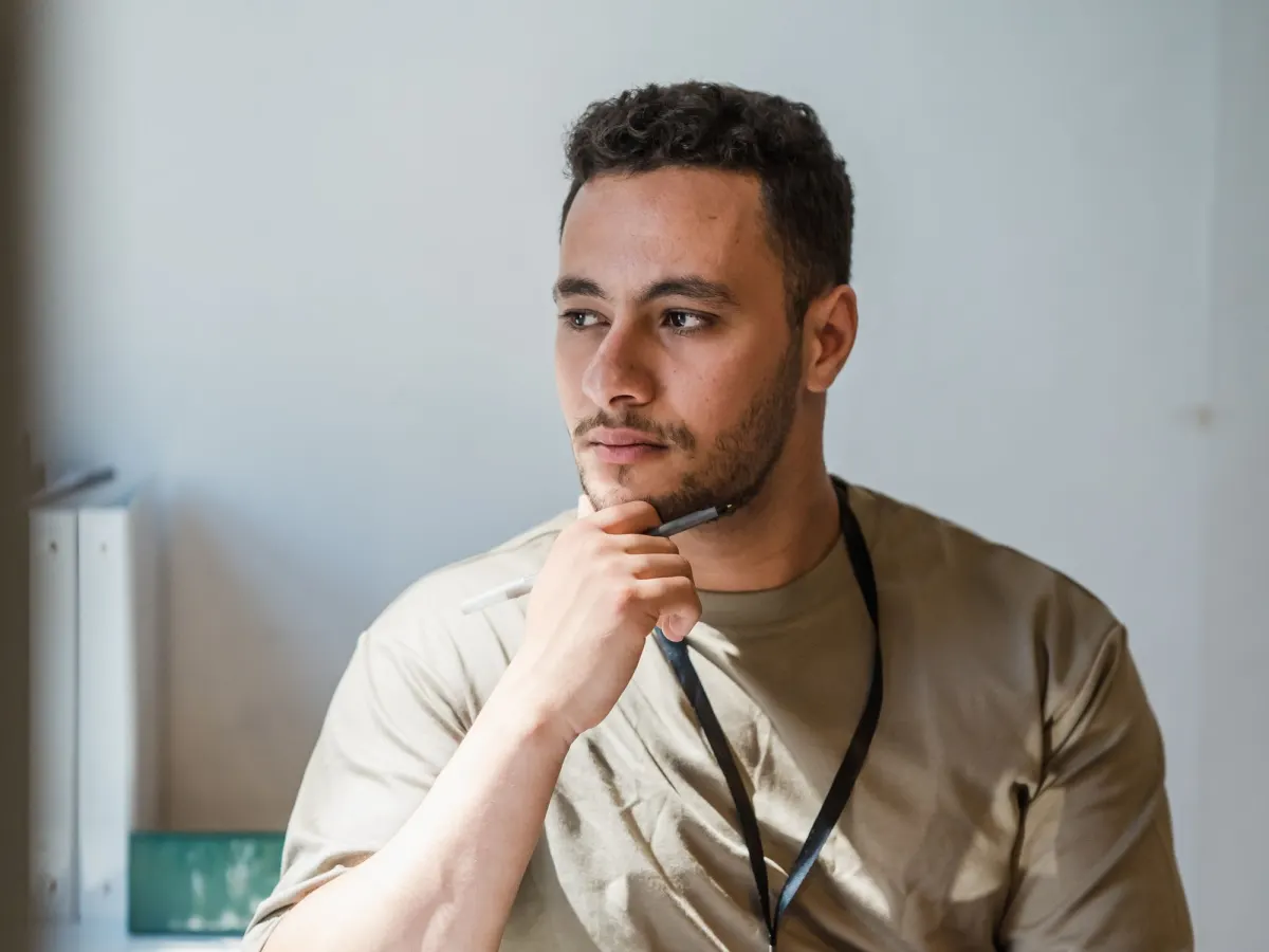 A man with a work lanyard sits by a sunny window, pen at his chin, thinking