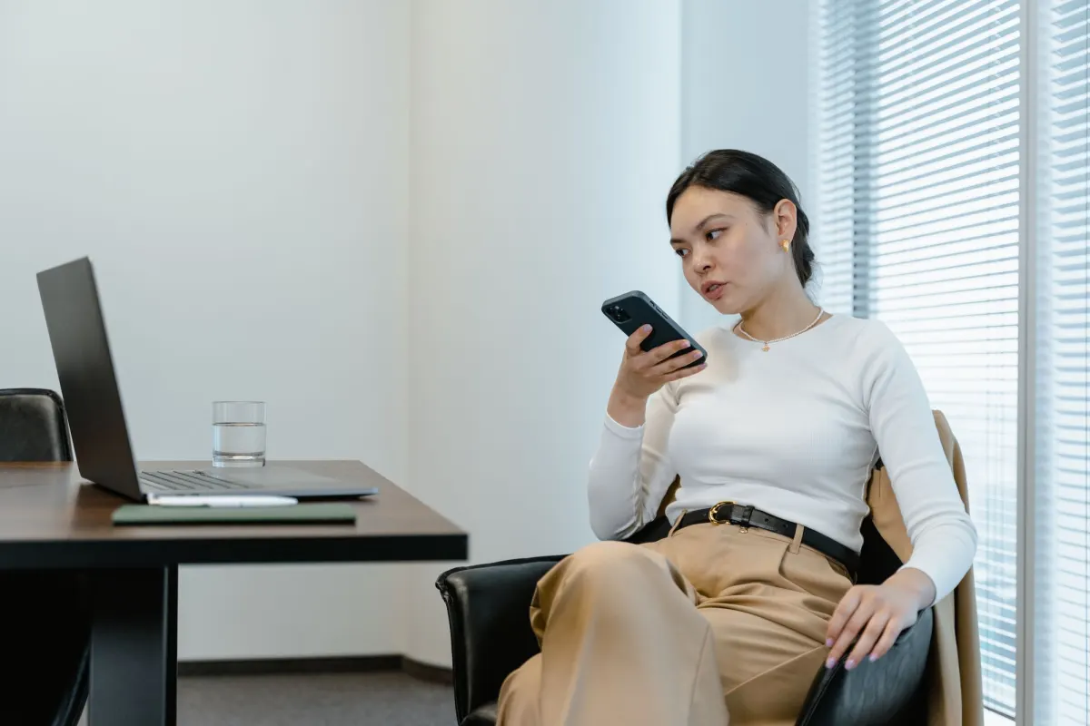 A woman sits alone in a quiet meeting room by the window blinds, speaking into her phone