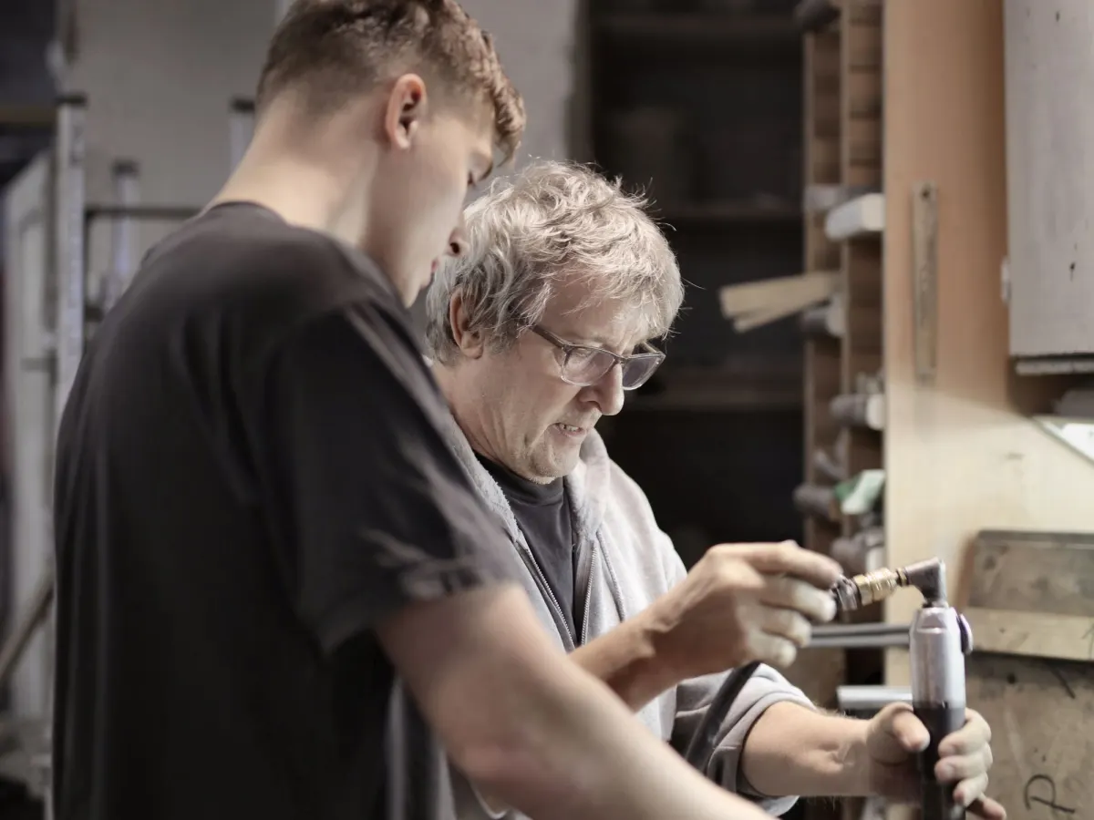 A young man in a dark T-shirt and an older man in glasses work side by side at a bench in a workshop