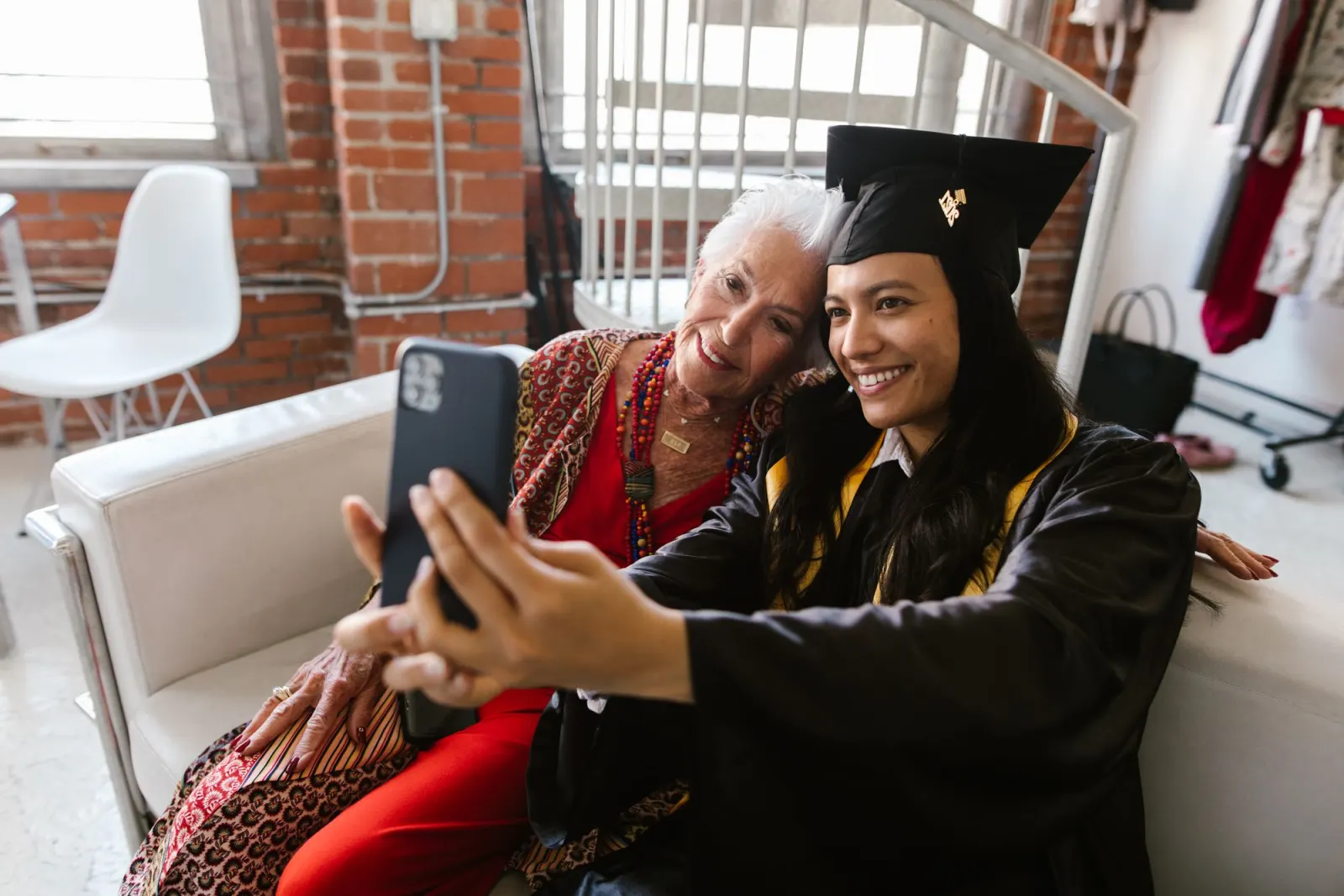 A smiling graduate in a black cap and gown takes a selfie with her white-haired grandmother on a sofa