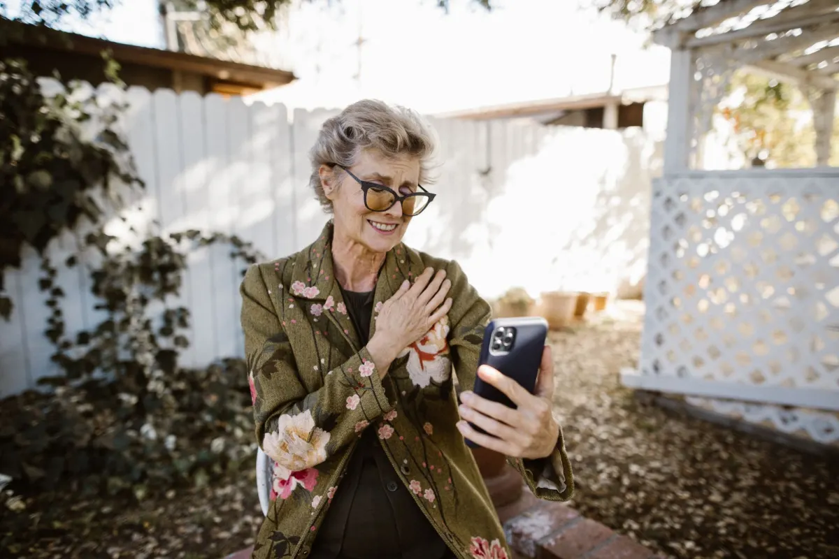 An older woman in a garden holds up her phone and speaks to it, one hand on her heart