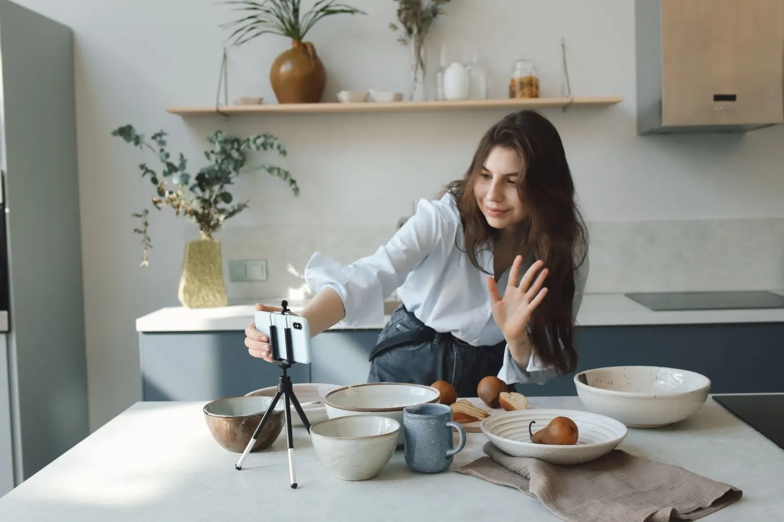 A woman waves at her phone, set on a small tripod on the kitchen bench, as she records a video