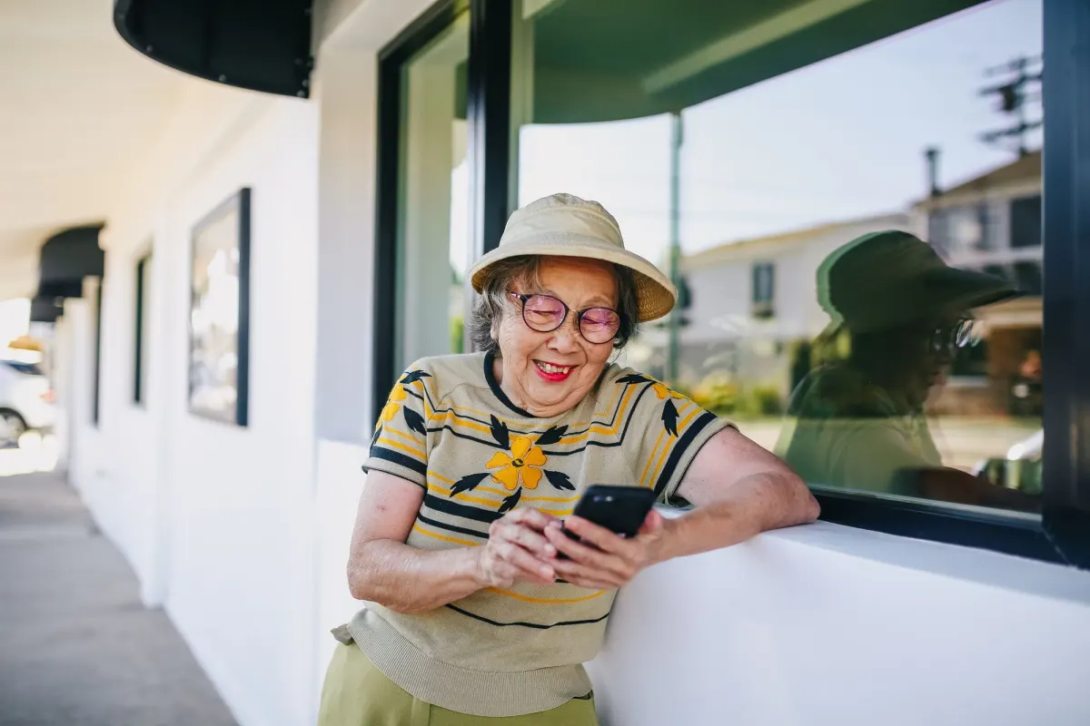 An older woman in a sun hat and glasses smiles at her phone outside a shop window