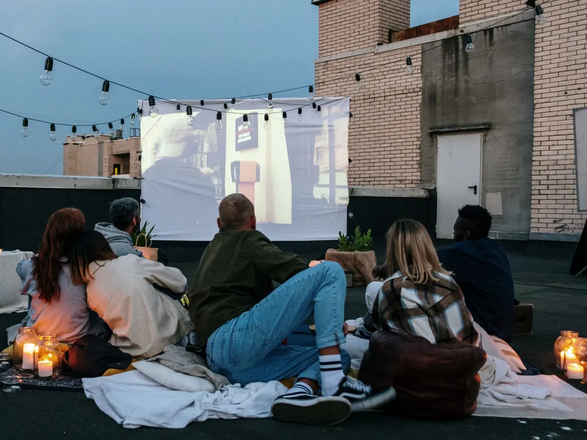 Friends sit on blankets on a rooftop at dusk, watching a film projected on a sheet under string lights