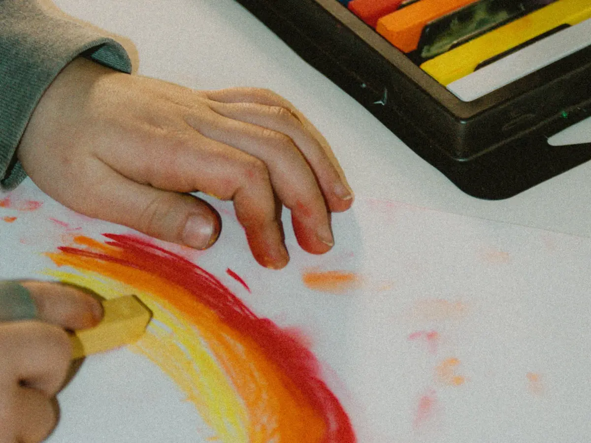 A small child's hands drawing a rainbow in red, orange and yellow pastels beside an open tray of pastels