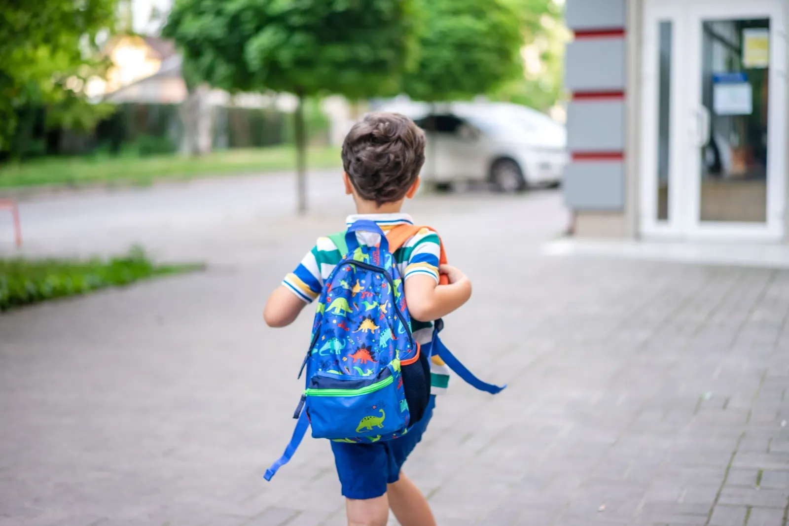 A young child in a striped shirt walks along a footpath wearing a blue dinosaur backpack, seen from behind