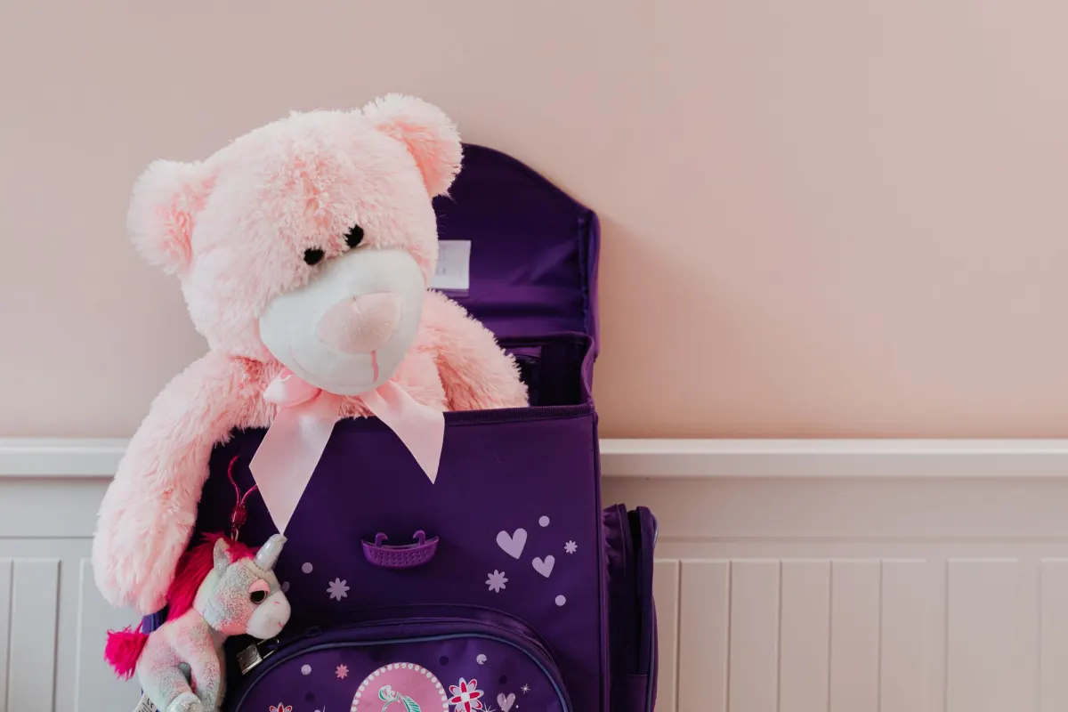 A pink teddy bear and a small unicorn toy tucked into an open purple school backpack against a pale wall