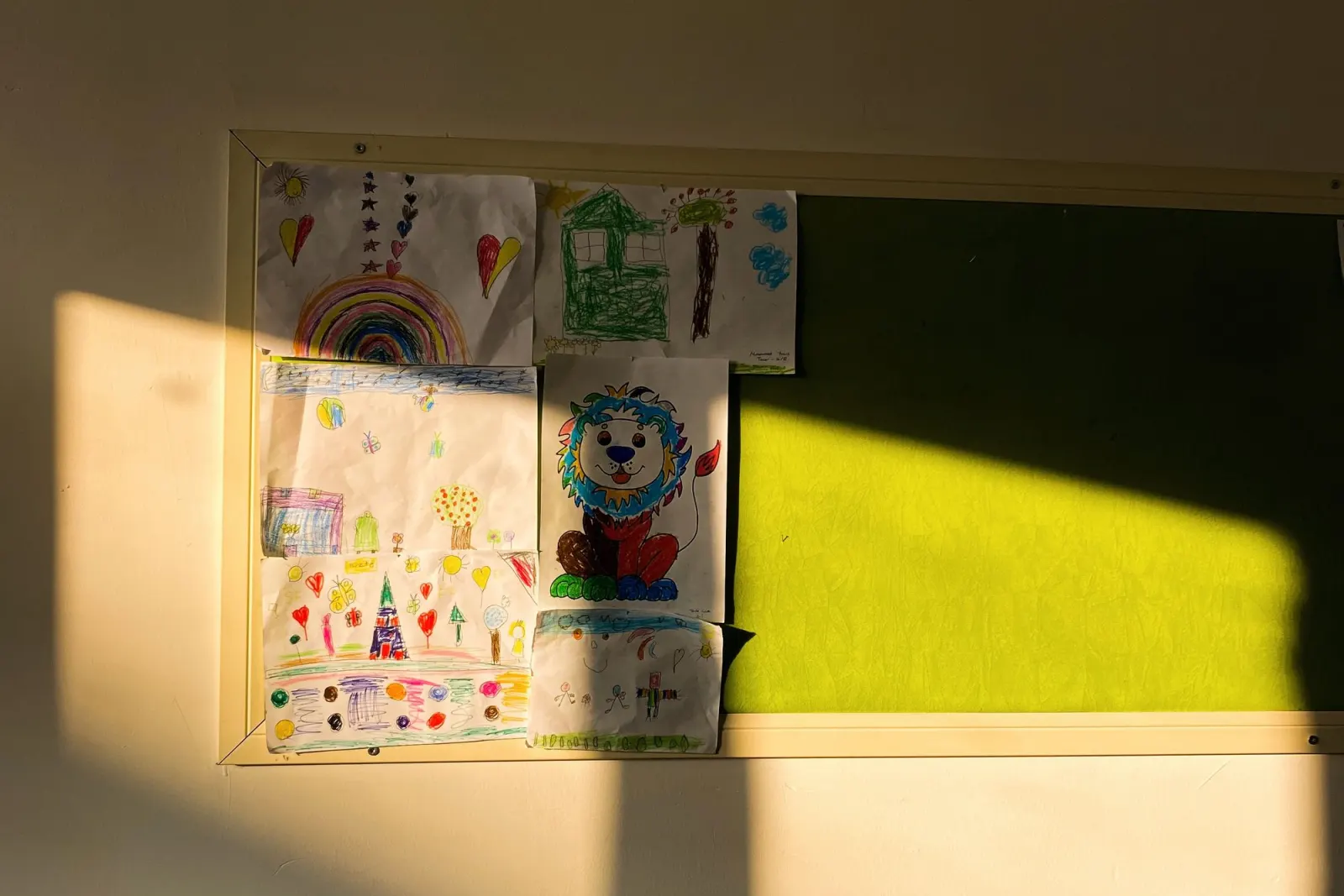 Children's crayon drawings of a rainbow, a house and a lion pinned to a classroom board in late afternoon sun