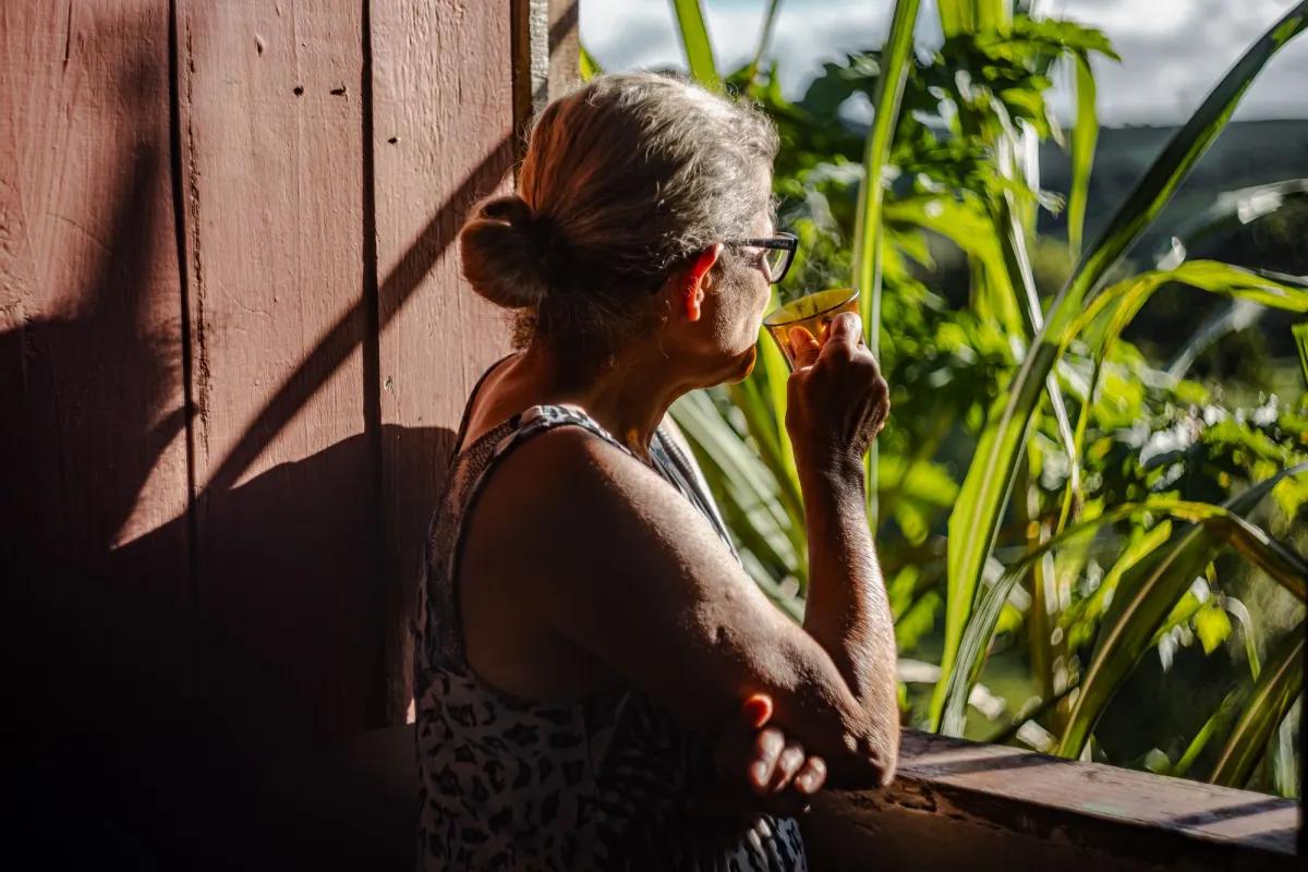 Older woman with grey hair sipping from a cup on a sunny verandah beside green plants