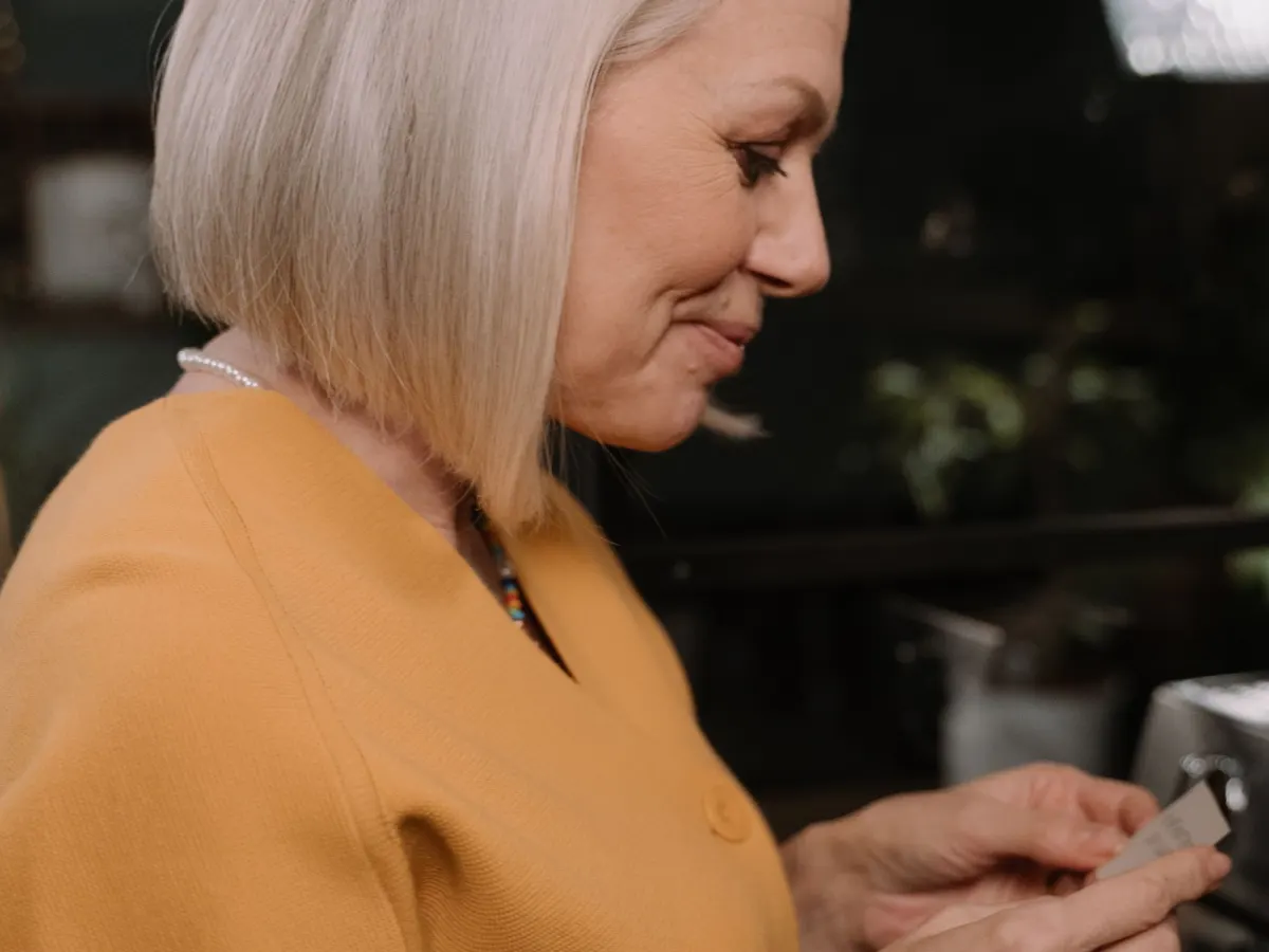 Woman with white hair and a yellow cardigan smiling as she reads a small card indoors