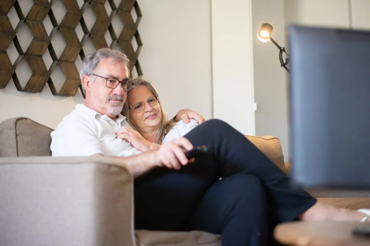 An older couple sit close together on a sofa, watching television at home