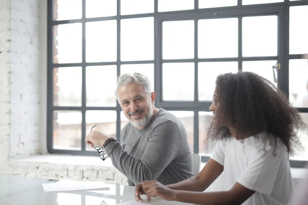 A grey-haired man smiles at a younger colleague across a table beside a large office window