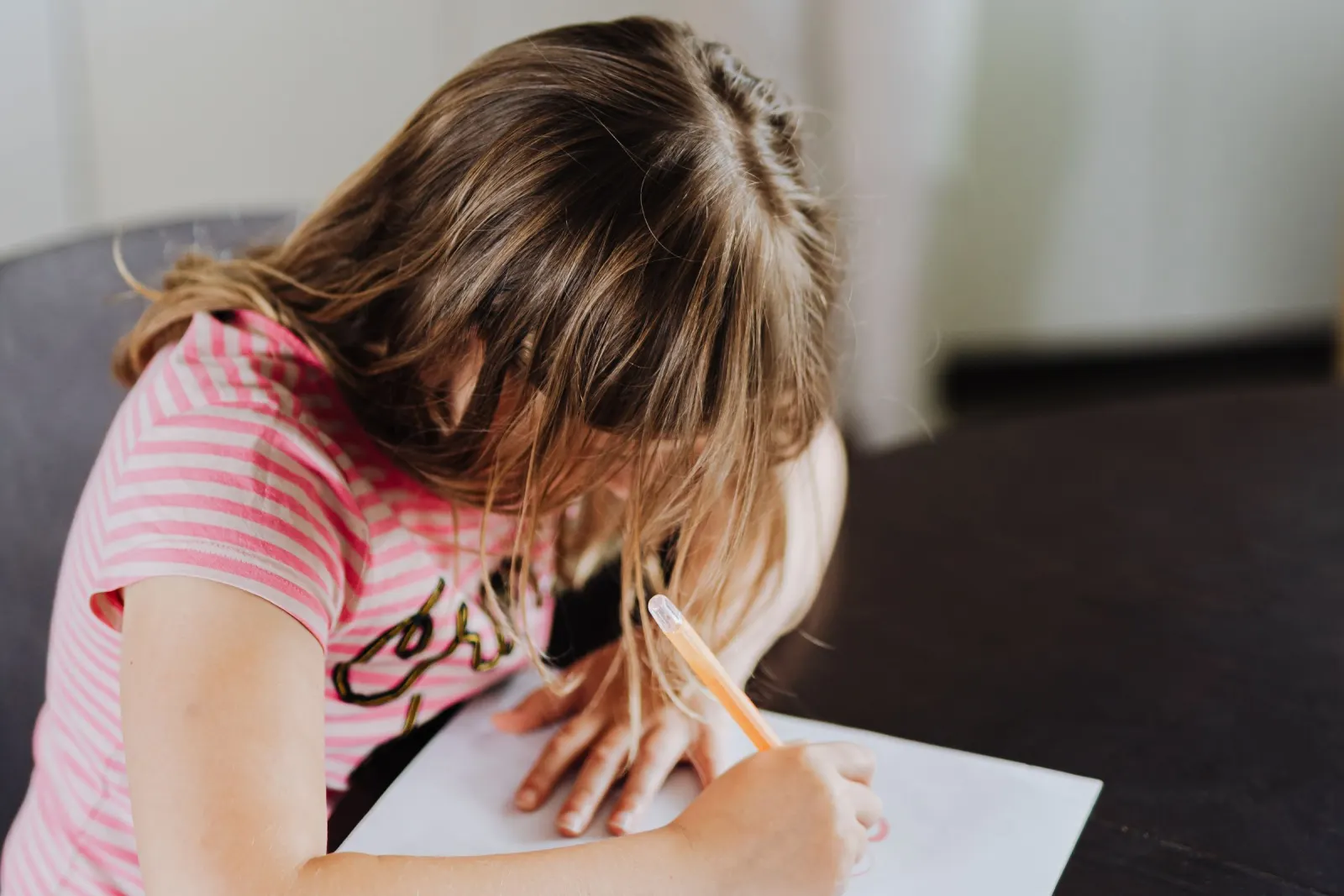 A young girl in a pink striped T-shirt leans over a sheet of paper at a dark table, writing with an orange pen