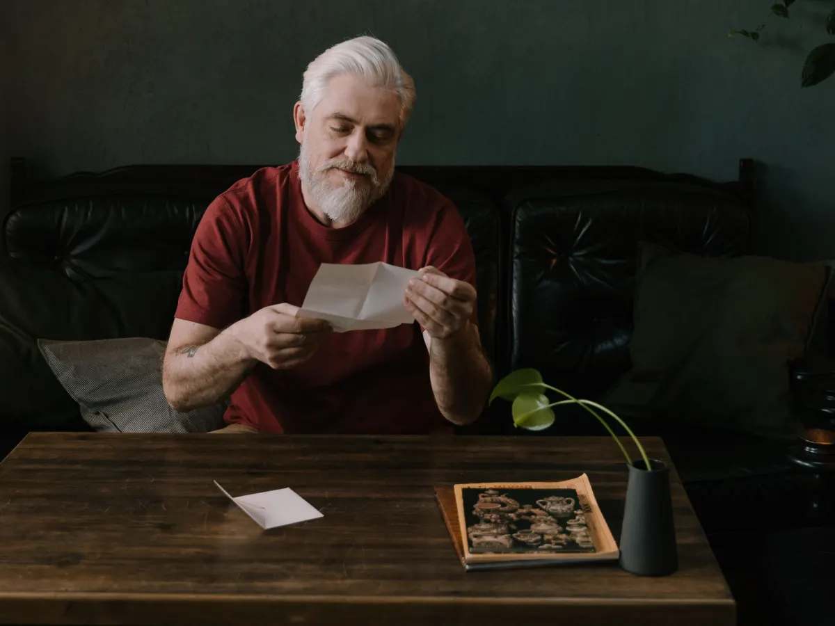 An older man with white hair and a beard reads a folded letter on a leather sofa at home