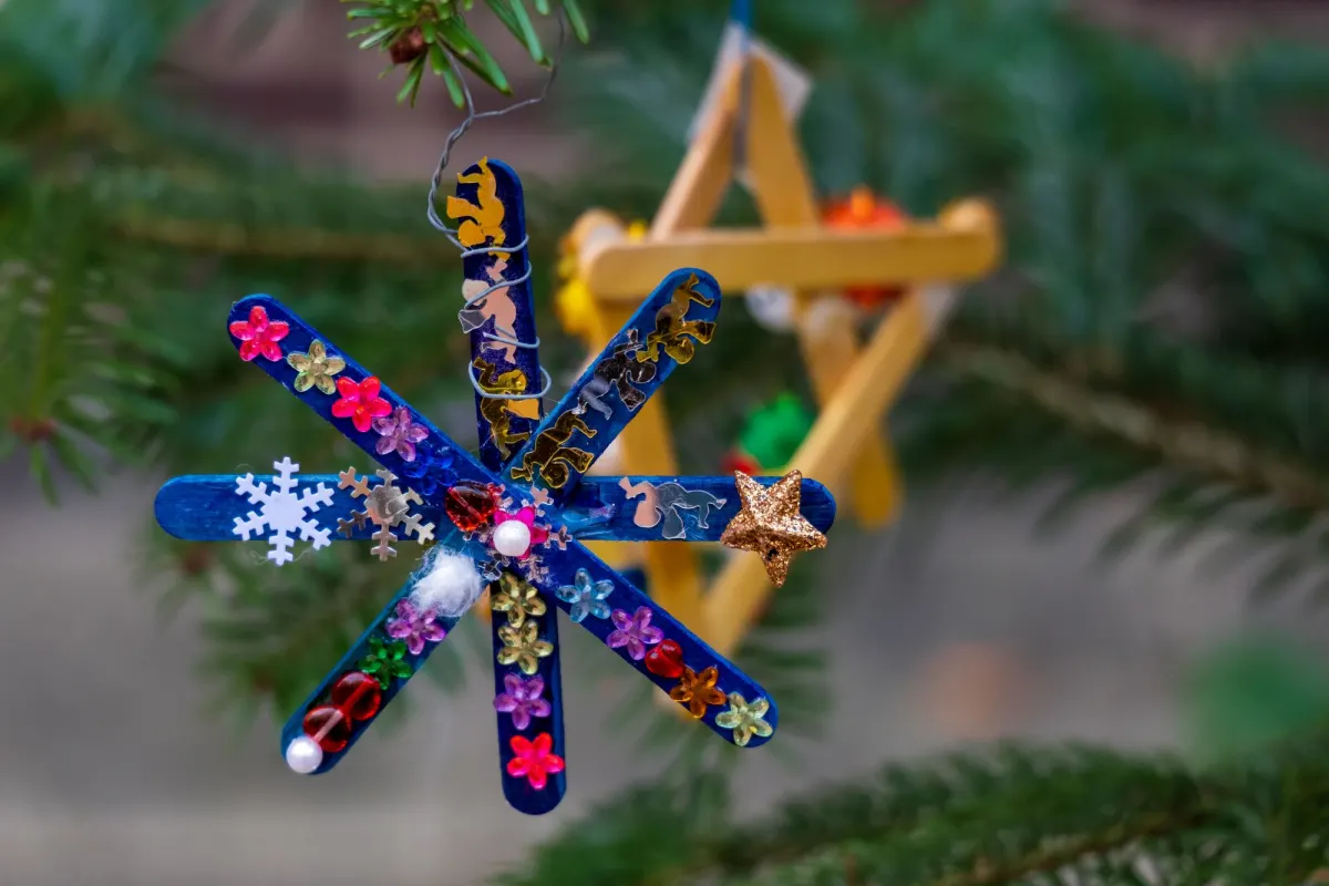A handmade star of blue paddle-pop sticks decorated with sequins, hanging on a Christmas tree in front of a yellow wooden star