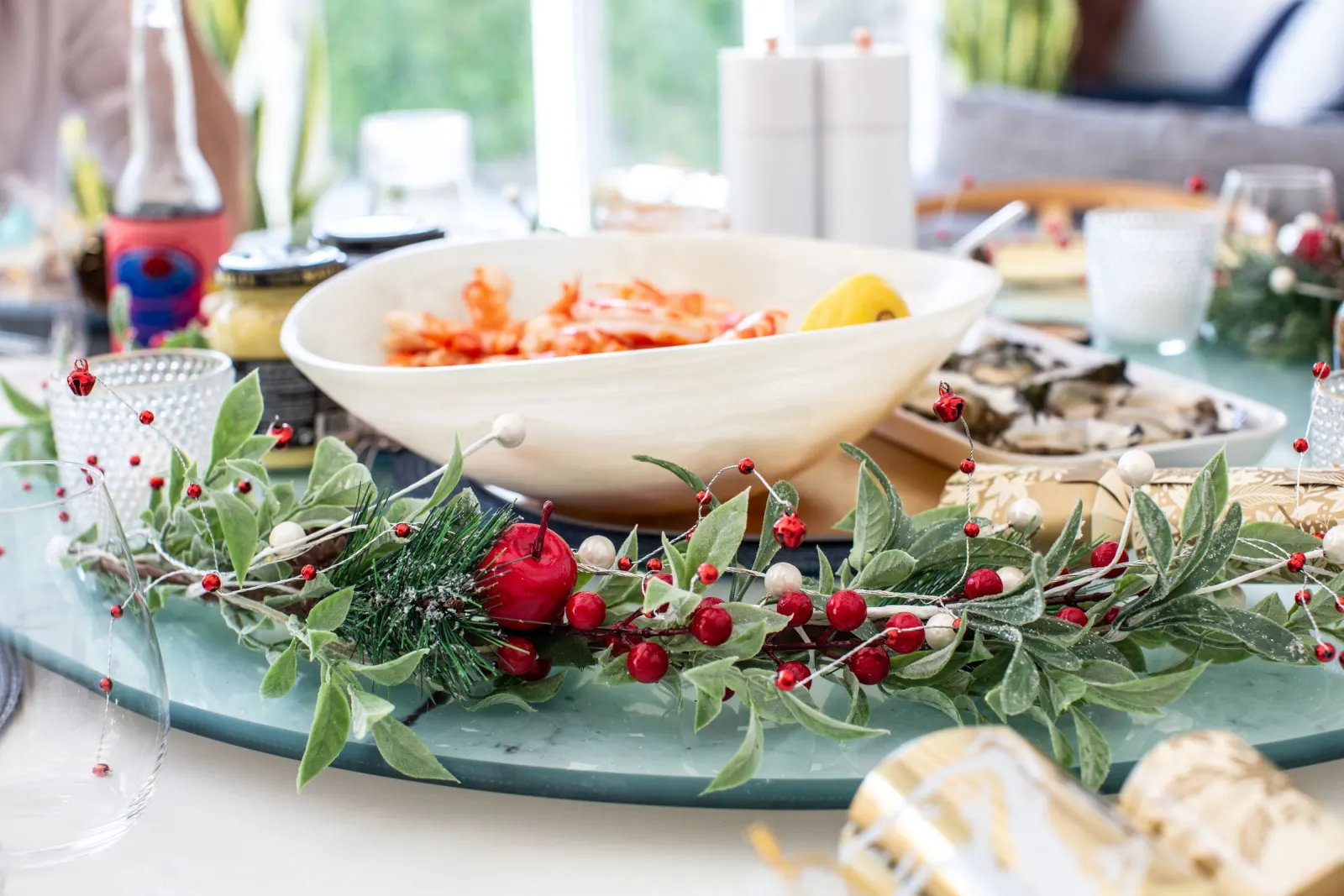 A Christmas lunch table with a bowl of prawns and a lemon, a plate of oysters, a red-berry garland and gold bonbons
