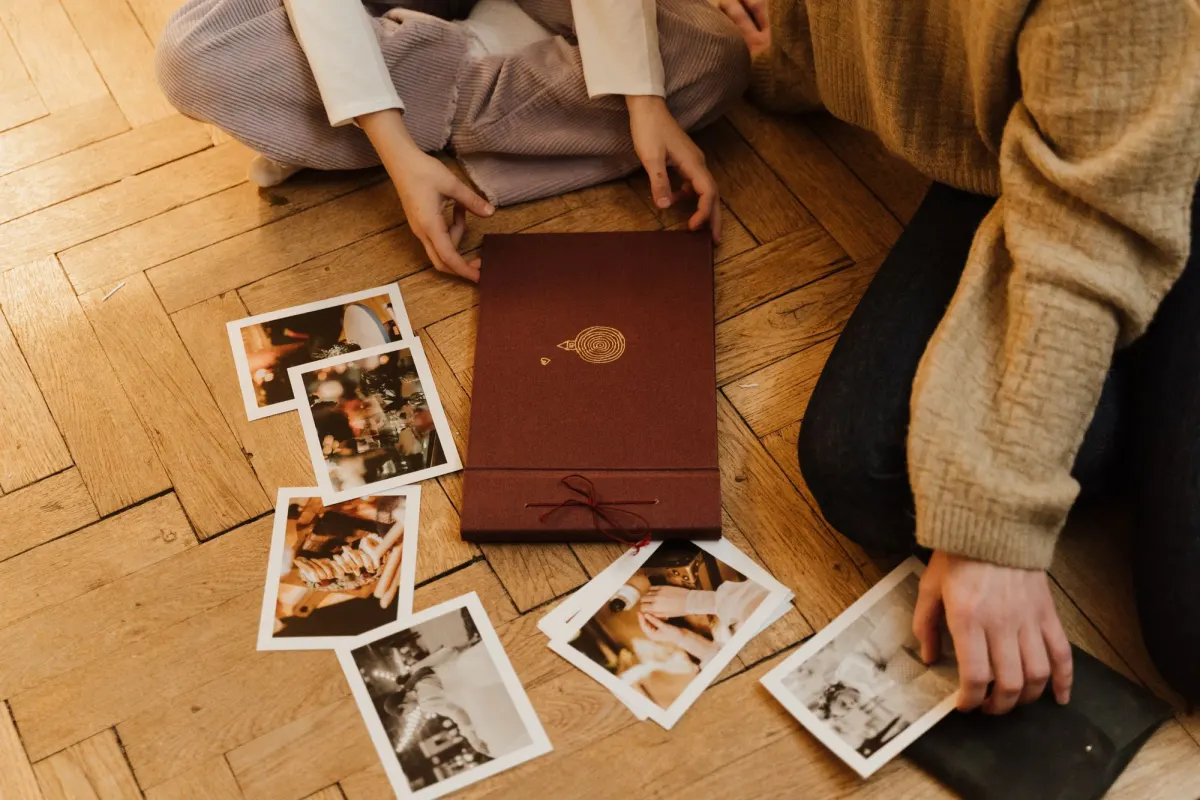 Printed photos spread on a wooden floor around a closed burgundy photo album, with hands sorting them