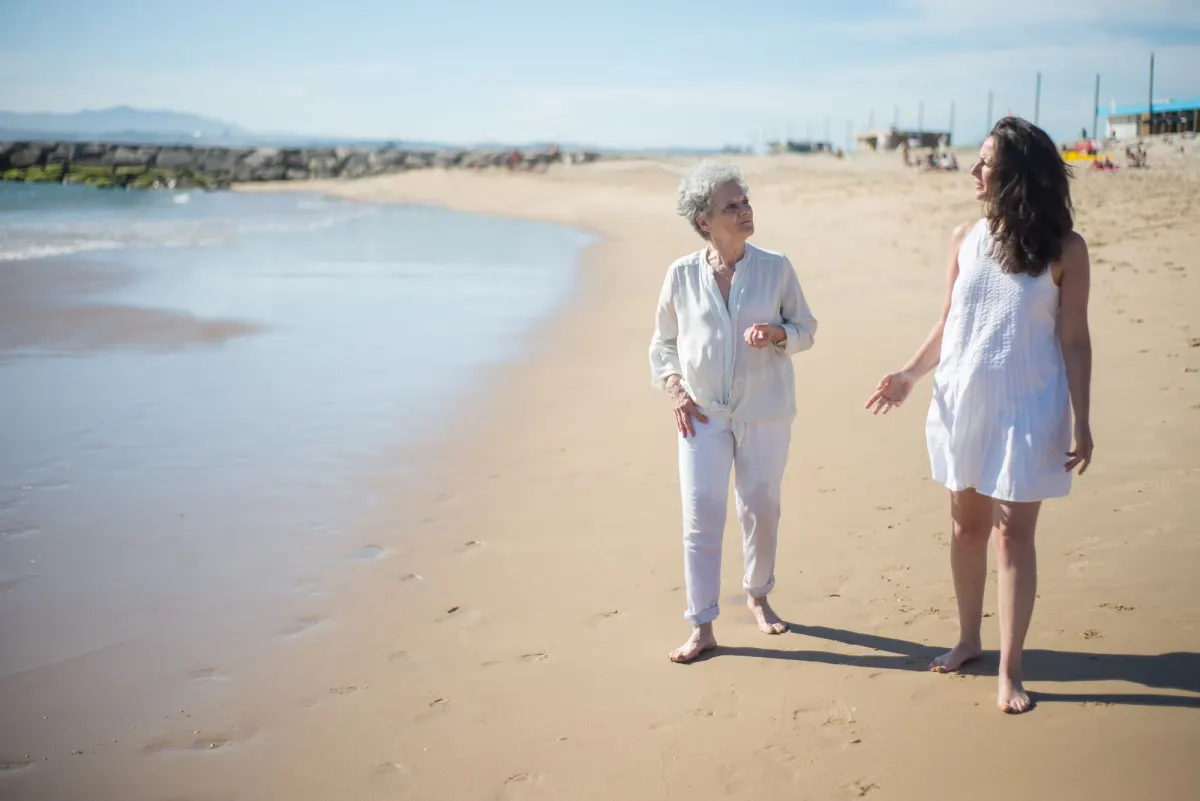 An older woman and a younger woman walk barefoot along a sunny beach, talking