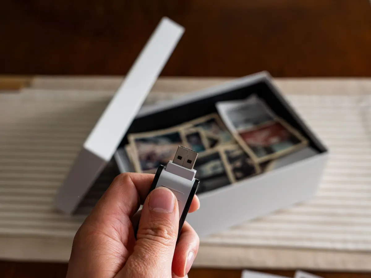 A hand holds a USB stick above an open white box of old family photos on a table