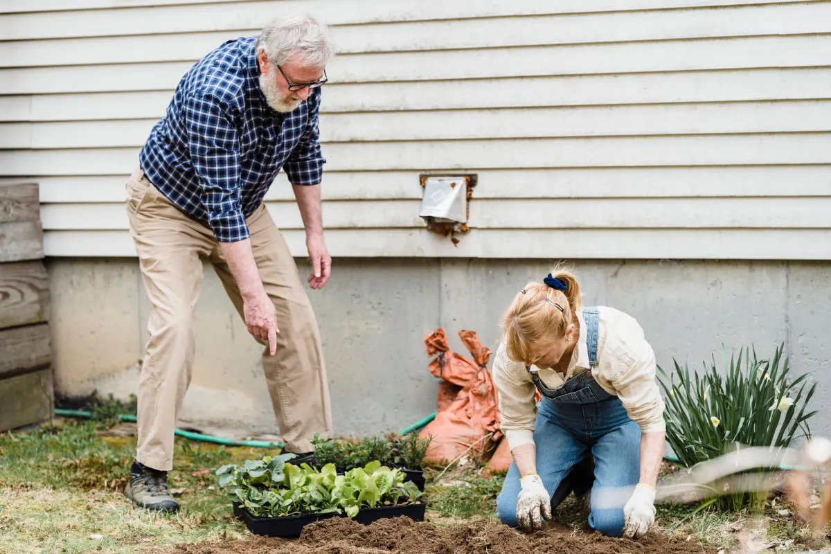 An older man in a checked shirt watches a woman in overalls plant seedlings beside a weatherboard house