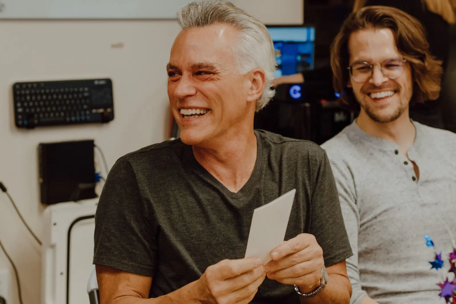 A grey-haired man laughs as he holds a small card at a workplace celebration, with a smiling younger colleague beside him