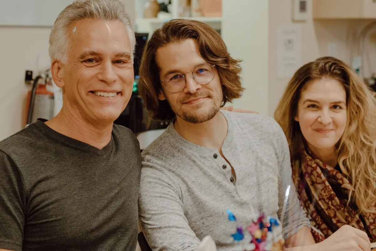 A grey-haired man and two younger colleagues sit close together and smile at the camera at a workplace celebration