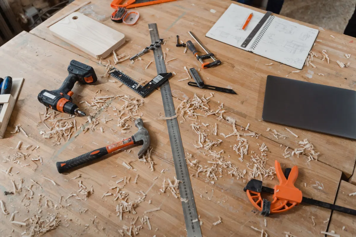 A cordless drill, hammer, steel rule, clamps and a sketchbook on a wooden workbench covered in wood shavings