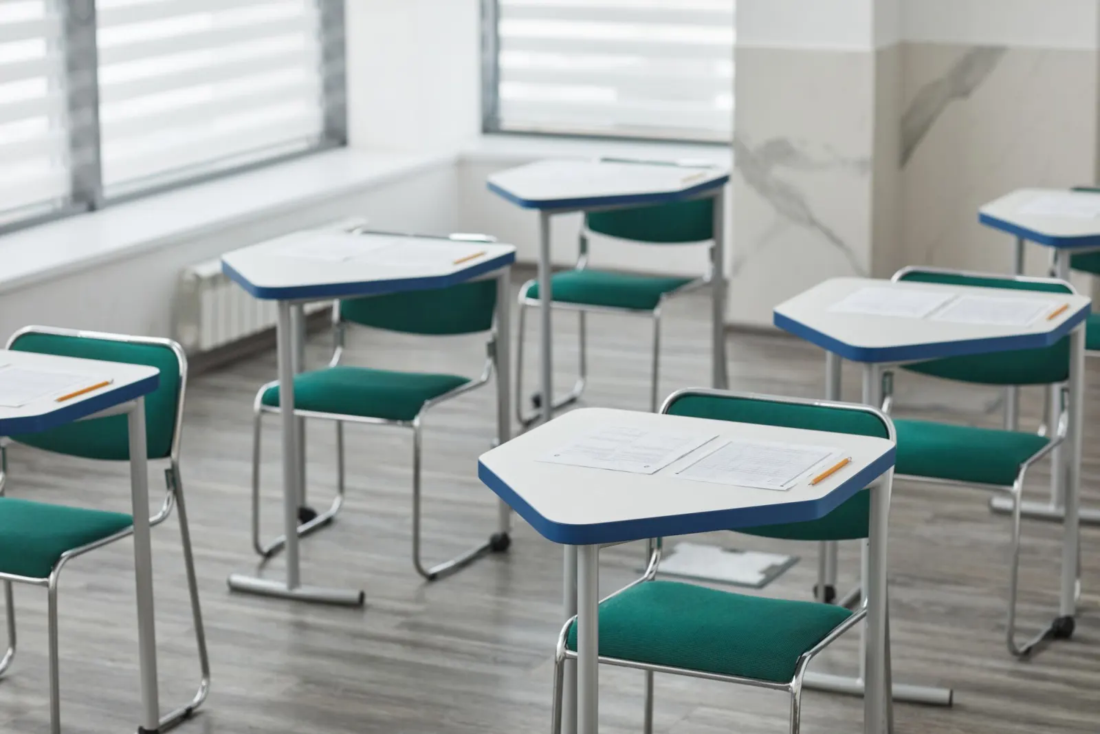 Empty exam desks with papers and pencils left on top, green chairs and soft daylight through window blinds