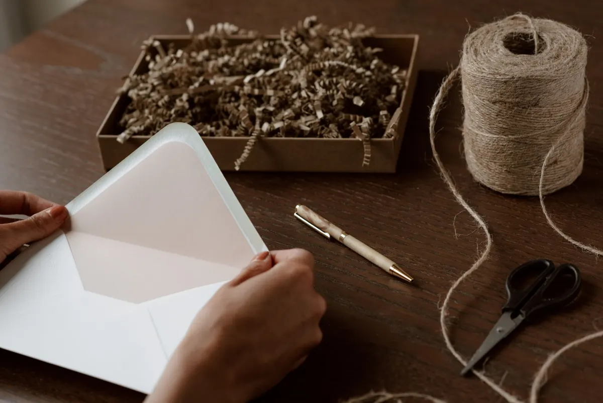 Hands open a pale pink envelope on a wooden desk beside a gift box, a pen, twine and scissors